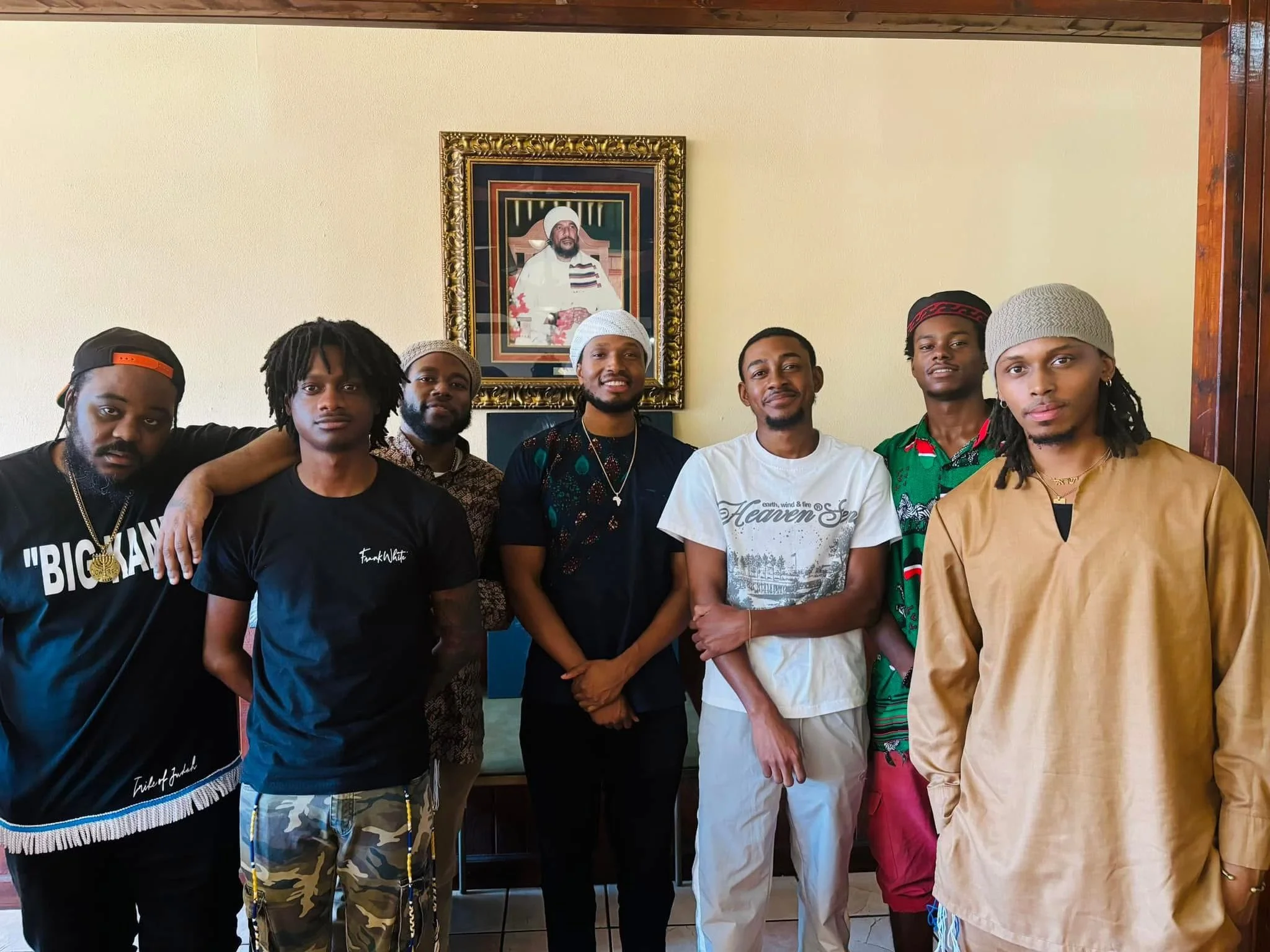 A group of seven young men (Israelites) standing in front of a portrait of a man in a frame on a beige wall. New Legacy Initiative. New Legacy, Inc.