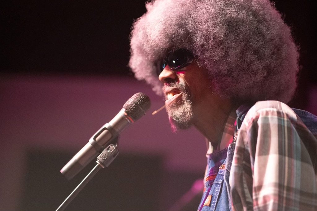 An Israelite man with large gray afro, sunglasses, and plaid shirt singing into a microphone on stage. New Legacy Initiative. New Legacy, Inc.