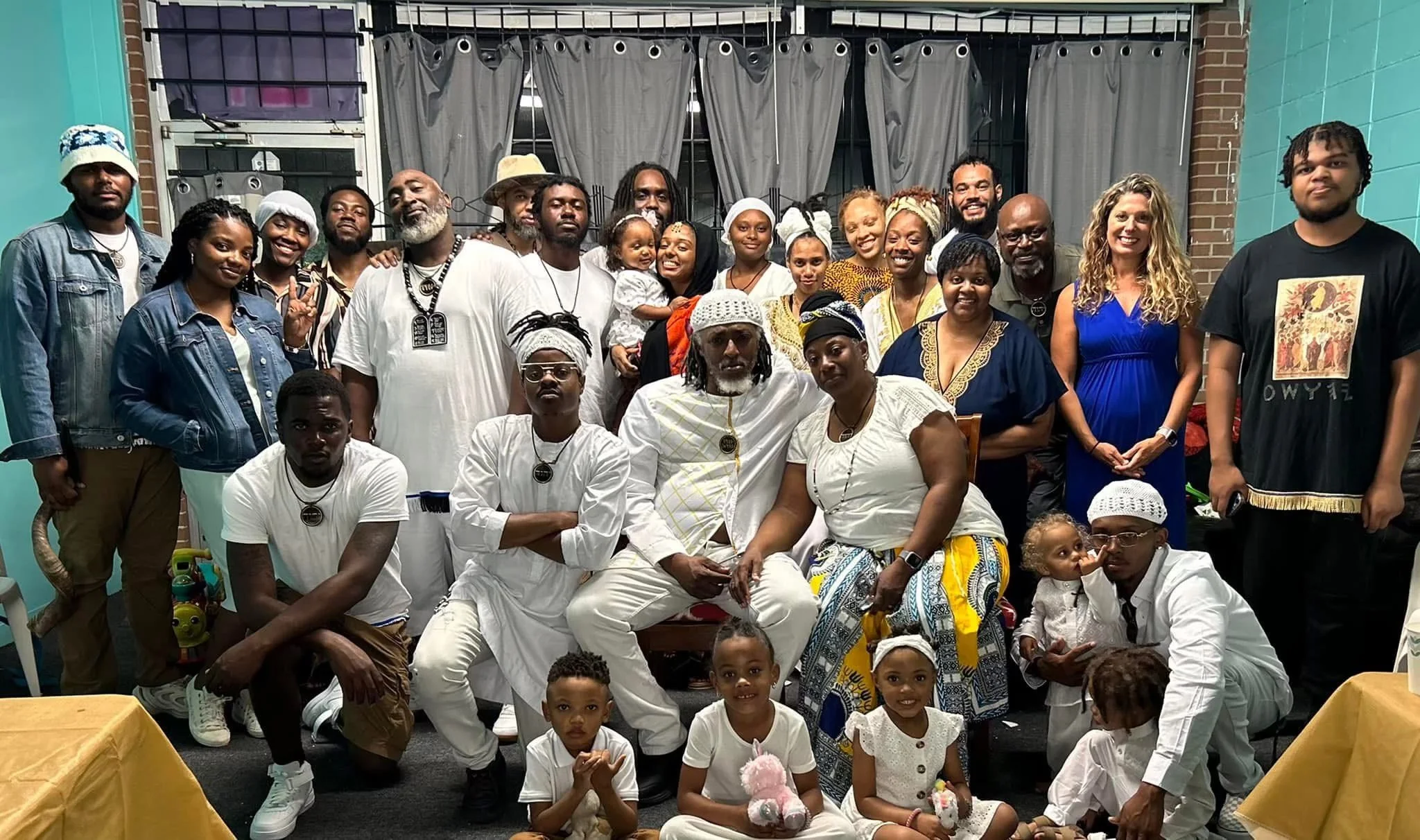 A diverse group of people (Israelites), including adults, children, and babies, gathered indoors in front of gray curtains, some dressed in white or traditional clothing, posing for a group photo. New Legacy Initiative. New Legacy, Inc.