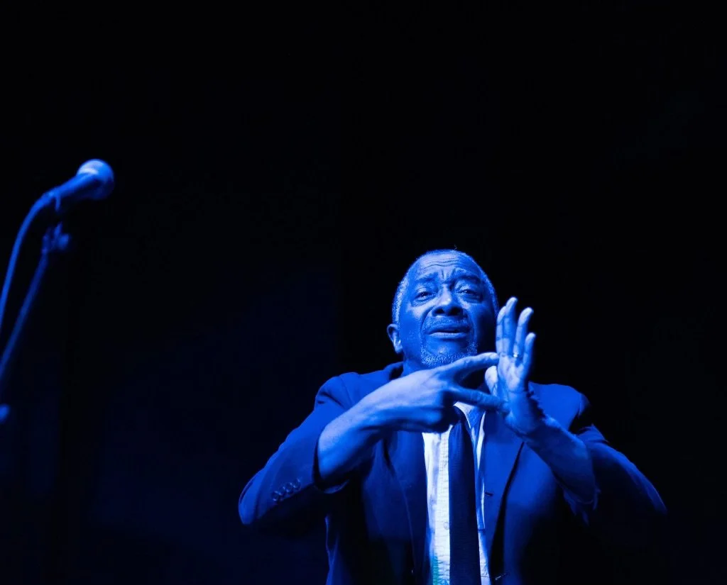 An Israelite  man in a suit performing on stage with blue lighting, standing in front of a microphone. New Legacy Initiative. New Legacy, Inc.