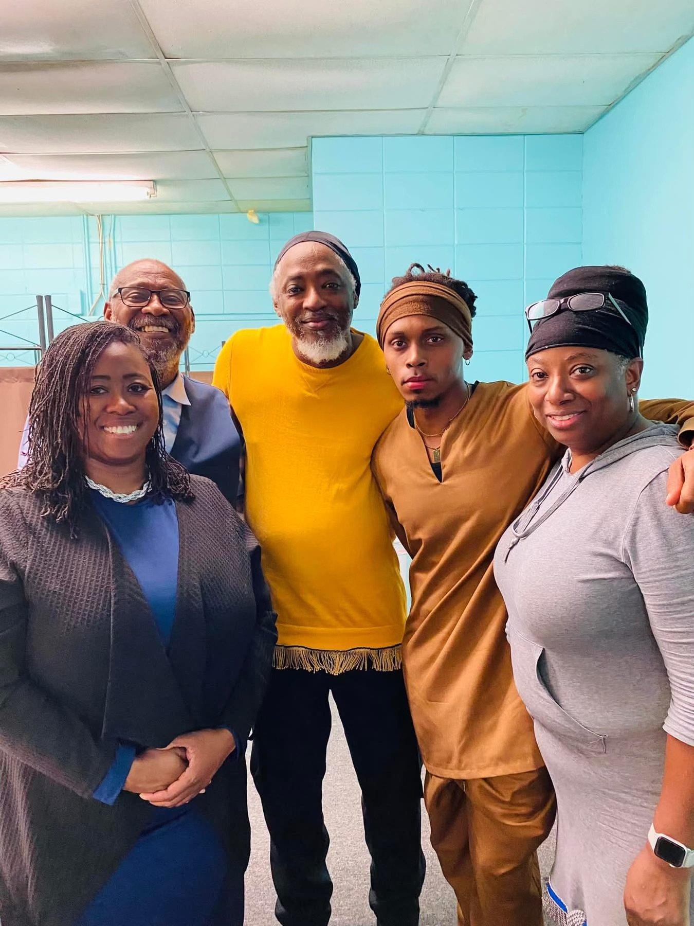 Group of six diverse people (Israelites) smiling and standing together indoors, with blue walls and ceiling tiles in the background. New Legacy Initiative. New Legacy, Inc.