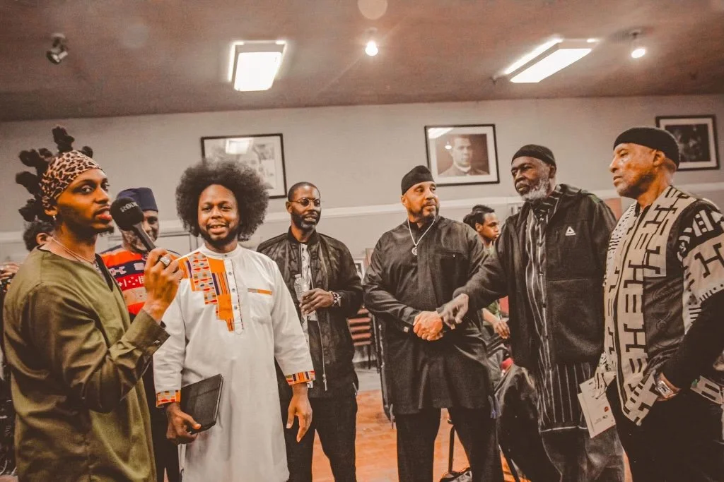 A group of men (Israelites), some shaking hands. One person on the left holding a microphone. Some individuals are dressed in traditional and modern clothing, and the overall scene appears to be a formal or social gathering. New Legacy, Inc.
