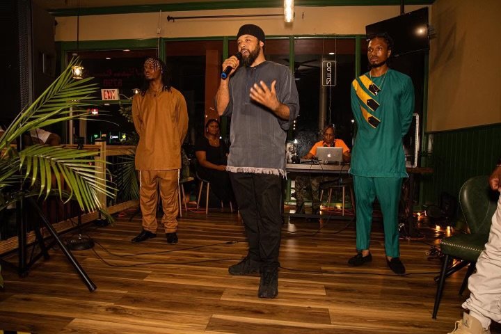 man with a beard and cap speaking into a microphone on a stage in a restaurant or bar, with two women standing behind him, one wearing a tan tracksuit and the other in a green outfit with yellow and black accents. Israelites. New Legacy, Inc.