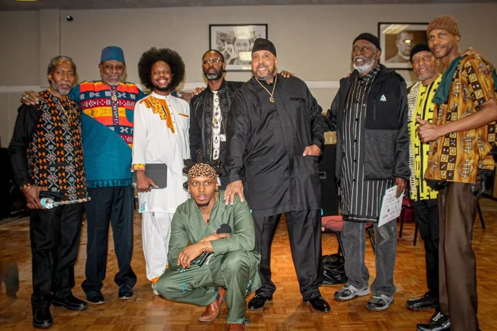A group of nine men (Israelites) posing for a photo indoors, some wearing traditional African clothing and others in casual or modern attire. New Legacy Initiative. New Legacy, Inc.