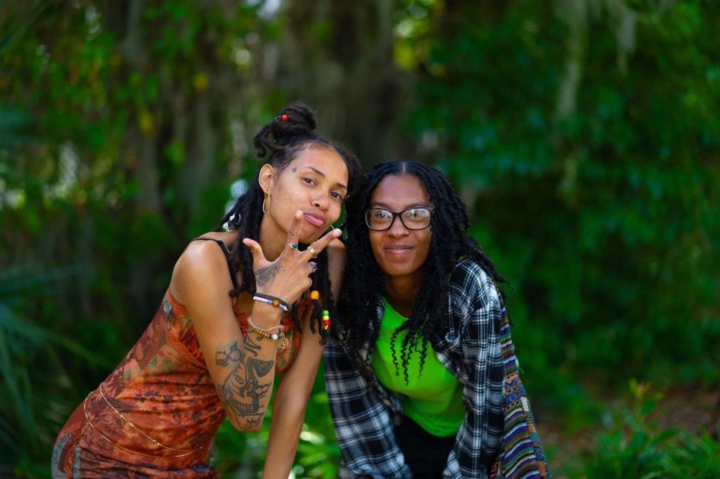 Two young (Israelite) women outdoors with green foliage background, one making a peace sign with hand, the other smiling. New Legacy Initiative. New Legacy, Inc.