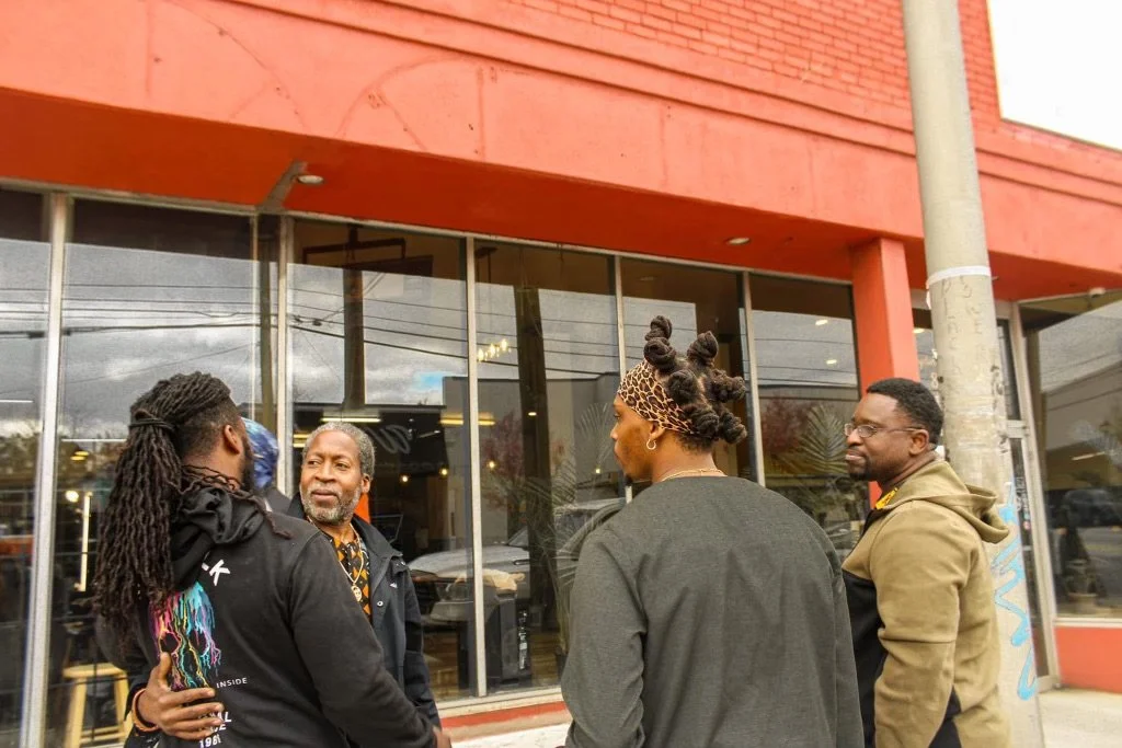 Four people (Israelites) are standing outside a building with large glass windows, engaging in conversation. New Legacy Initiative. New Legacy, Inc.