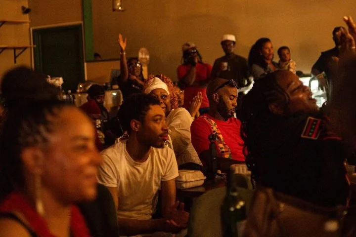 Israelites sitting at tables and standing in a dimly lit room, attending an event or gathering, some with hands raised. New Legacy Initiative. New Legacy, Inc.