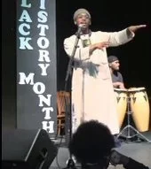 An Israelite woman in a white dress and headscarf singing on stage with a microphone, accompanied by a percussionist playing conga drums. New Legacy Initiative. New Legacy, Inc.