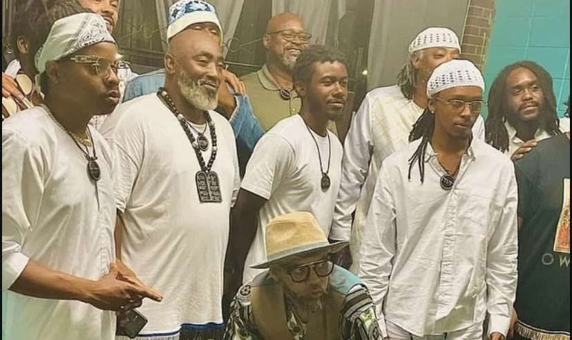 A group of men (Israelites) of diverse ages and appearances, some wearing head coverings and religious necklaces, are gathered together for a photo. One man in the front is seated with a hat, glasses, and a patterned shirt. New Legacy, Inc.