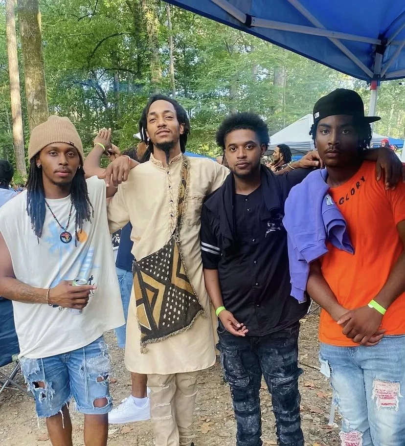 Four young men (Israelites) standing together outdoors under a blue canopy, with a forest background and other people in the distance. New Legacy Initiative. New Legacy, Inc.