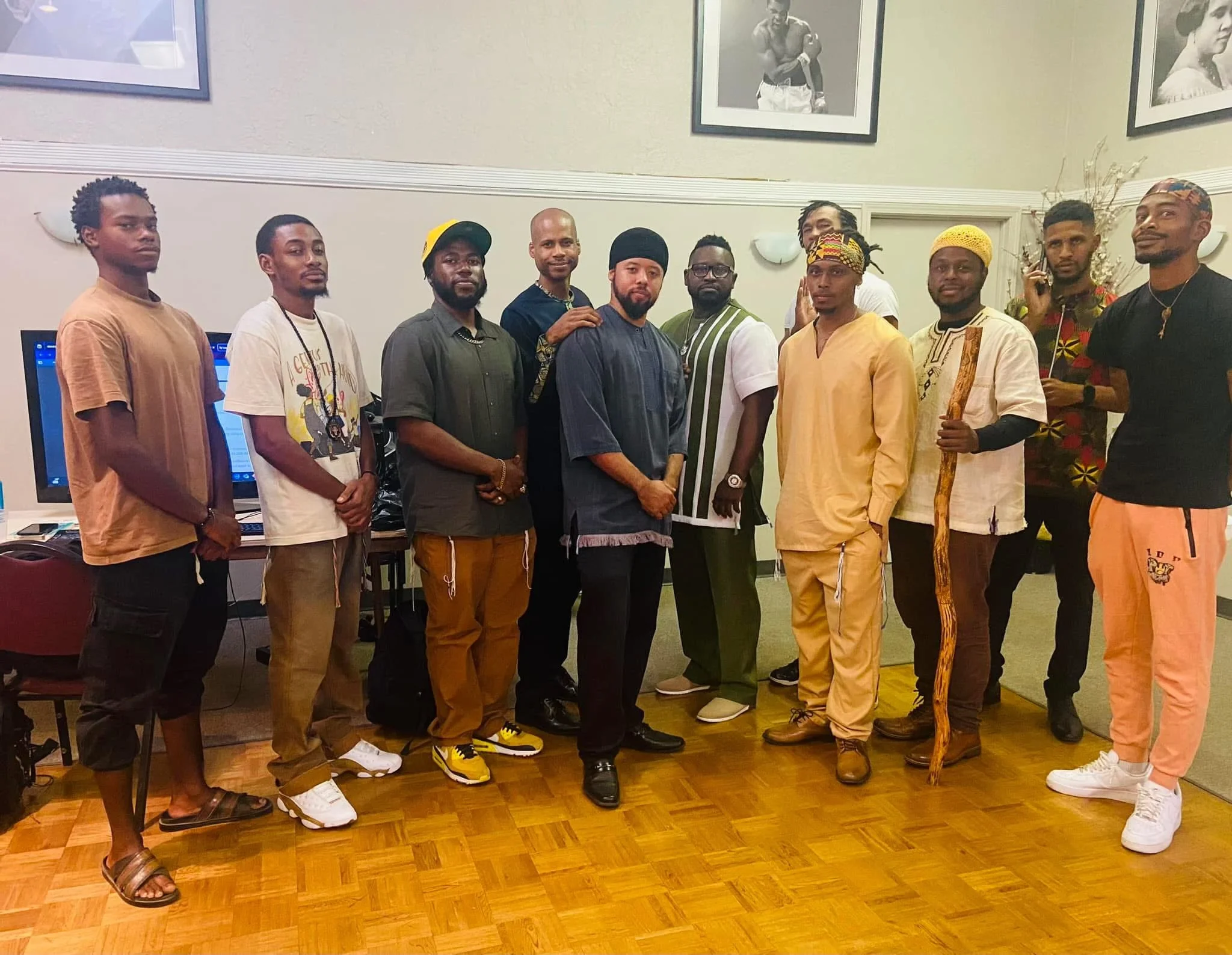 A group of eleven men (Israelites) standing side by side indoors, some in traditional African attire and others in casual clothing, with framed black and white photographs on the beige wall behind them. New Legacy Initiative. New Legacy, Inc.