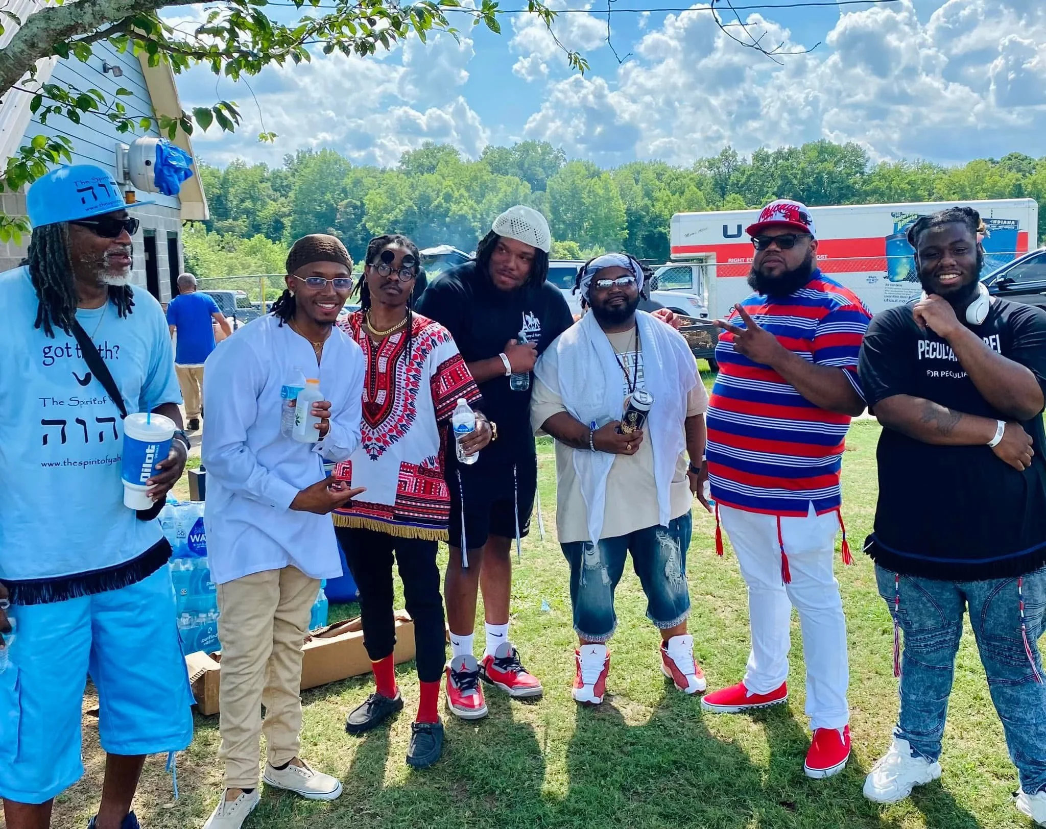 A group of eight men (Israelites) standing outdoors on a sunny day near a wooded area, with some holding water bottles and smiling. New Legacy Initiative. New Legacy, Inc.