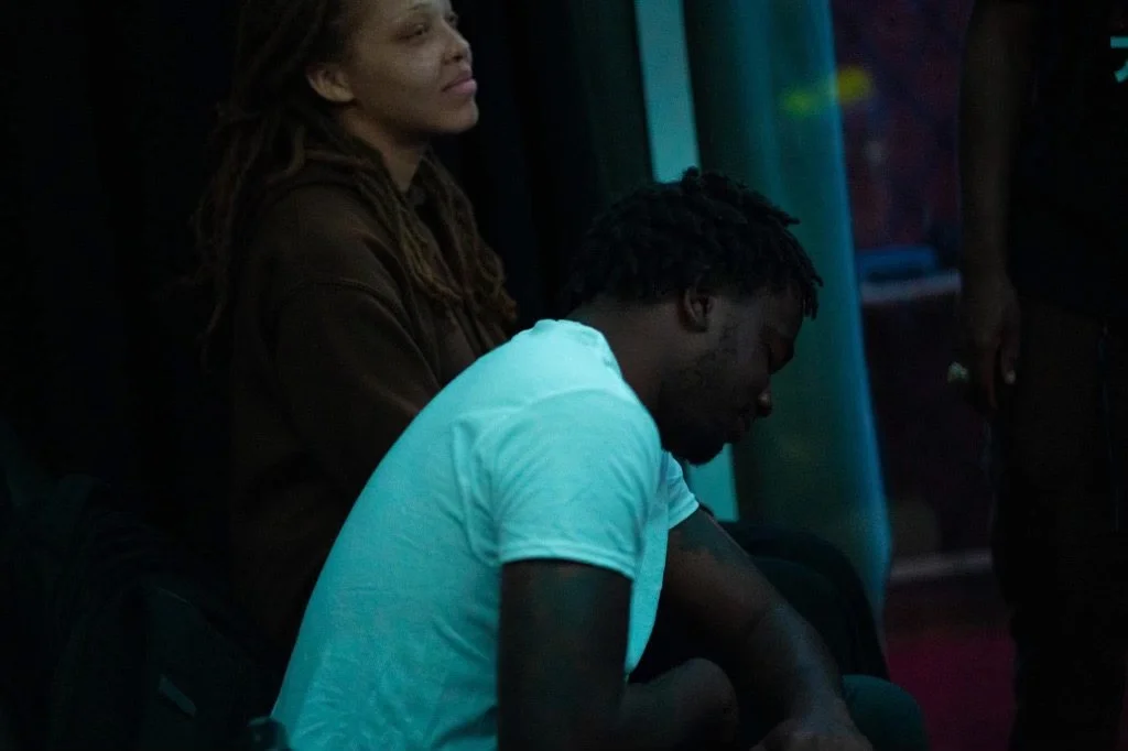 An Israelite woman with locs and a man with short dreadlocks sitting with their heads bowed in a dark room. New Legacy Initiative. New Legacy, Inc.