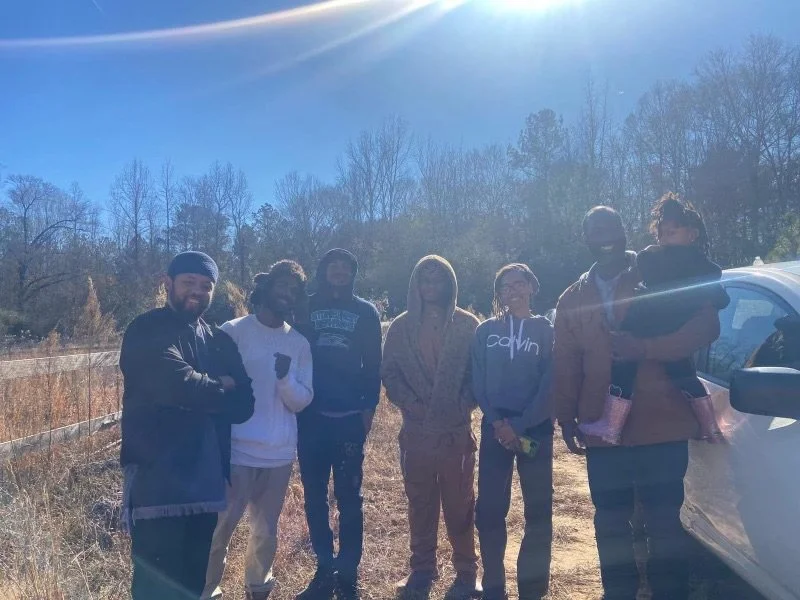Group of seven people (Israelites) standing outdoors on a sunny day, with trees and a car visible in the background. New Legacy Initiative. New Legacy, Inc.