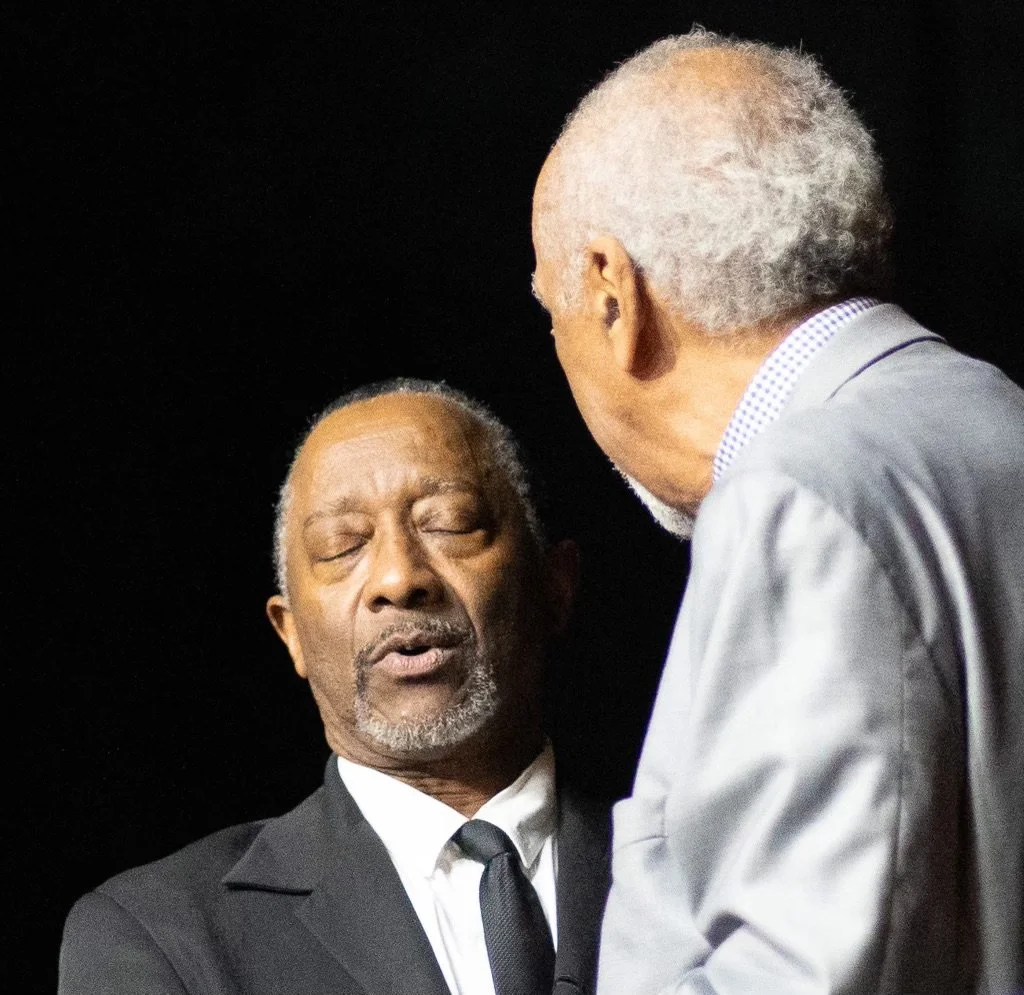 Two older men (Israelites) engaged in a serious conversation; one with closed eyes, wearing a black suit and tie, the other with gray hair and beard, dressed in a gray suit, against a dark background. New Legacy Initiative. New Legacy, Inc.