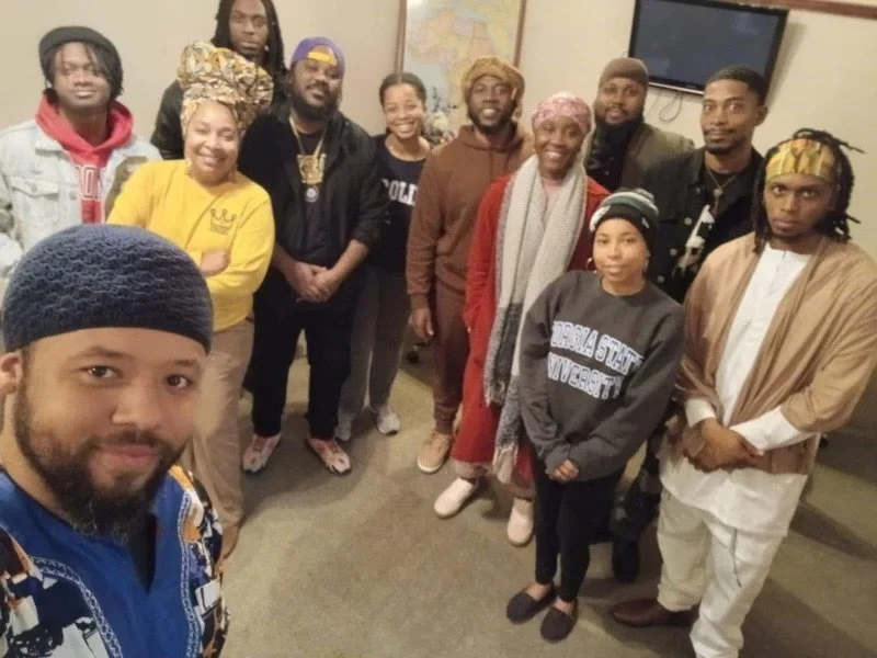 A diverse group of people posing in an indoor setting, smiling and looking at the camera. Some are wearing casual and colorful clothing, including headscarves and hats. A man in the foreground is taking a selfie.