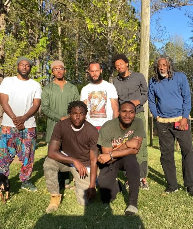 A group of eight men (Israelites) standing outdoors on grass, with trees and a utility pole in the background on a sunny day. New Legacy Initiative. New Legacy, Inc.