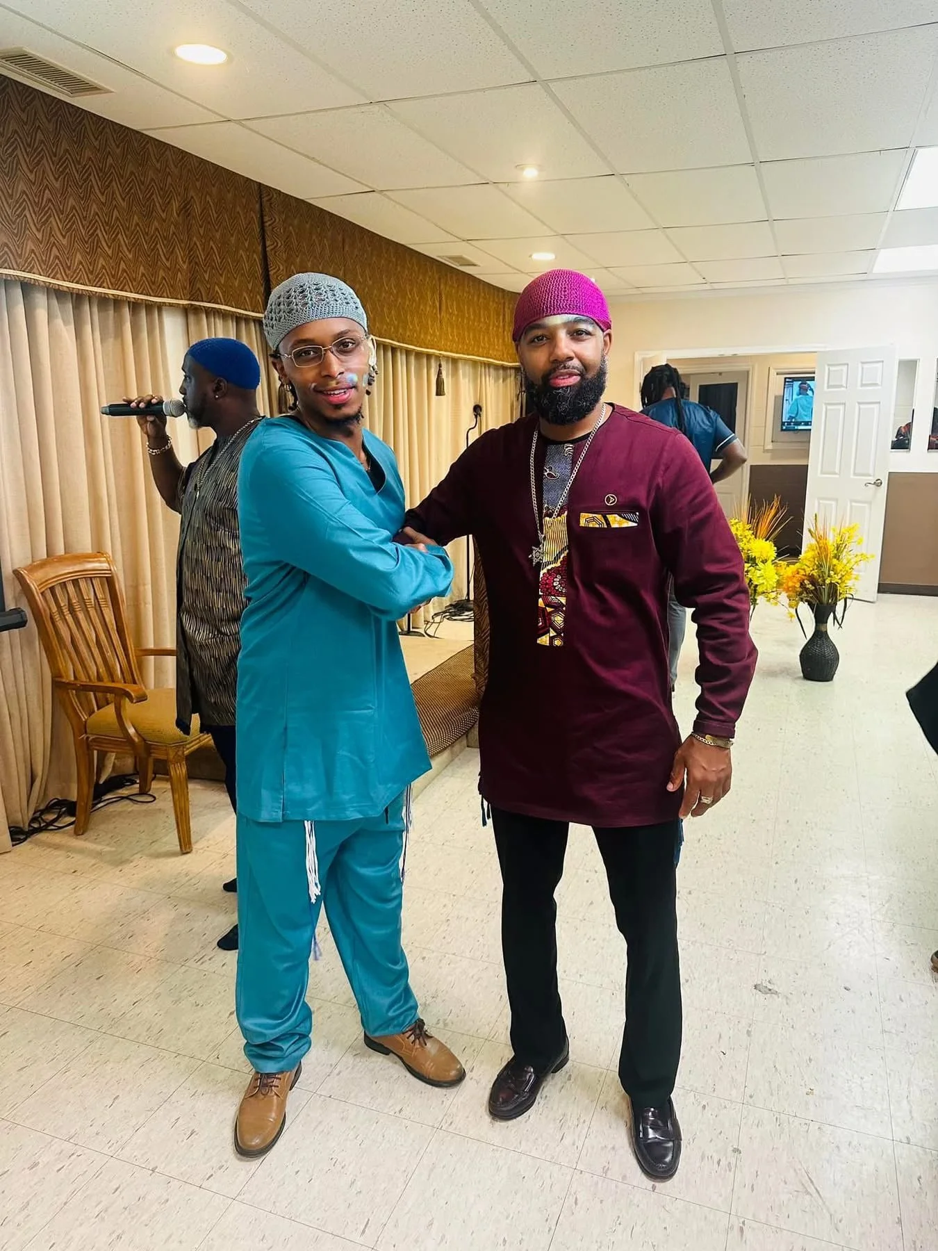 Two (Israelite) men standing in a room, shaking hands and smiling at the camera. One is dressed in blue scrubs and a matching cap, while the other is wearing a maroon shirt and a pink cap. New Legacy Initiative. New Legacy, Inc.