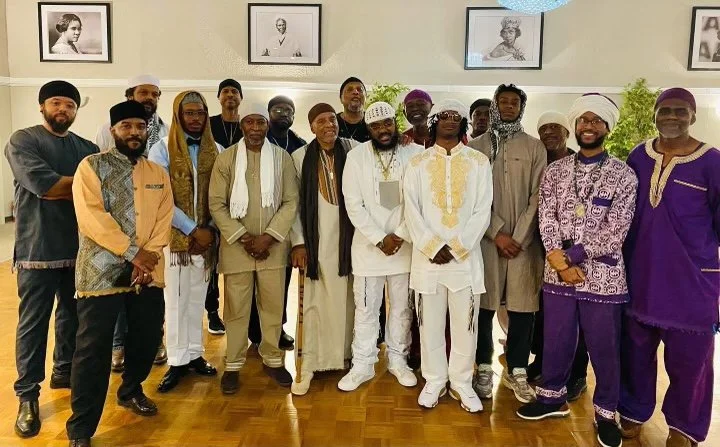 Group of men (Israelites) dressed in traditional African and modern clothing, standing indoors in front of a wall with framed black-and-white photographs. New Legacy Initiative. New Legacy, Inc.