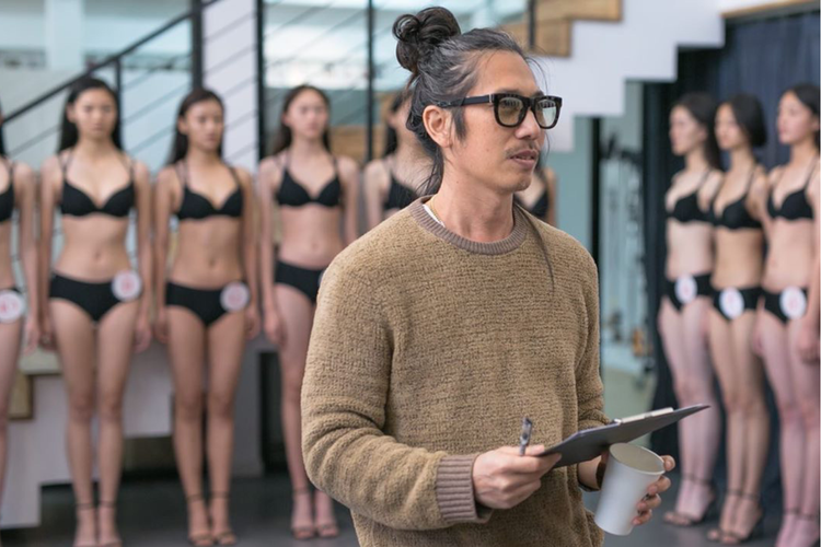 Man with glasses and a sweater holding a clipboard and a coffee cup, standing in front of a row of women in swimsuits, possibly contestants in a beauty pageant or modeling event.