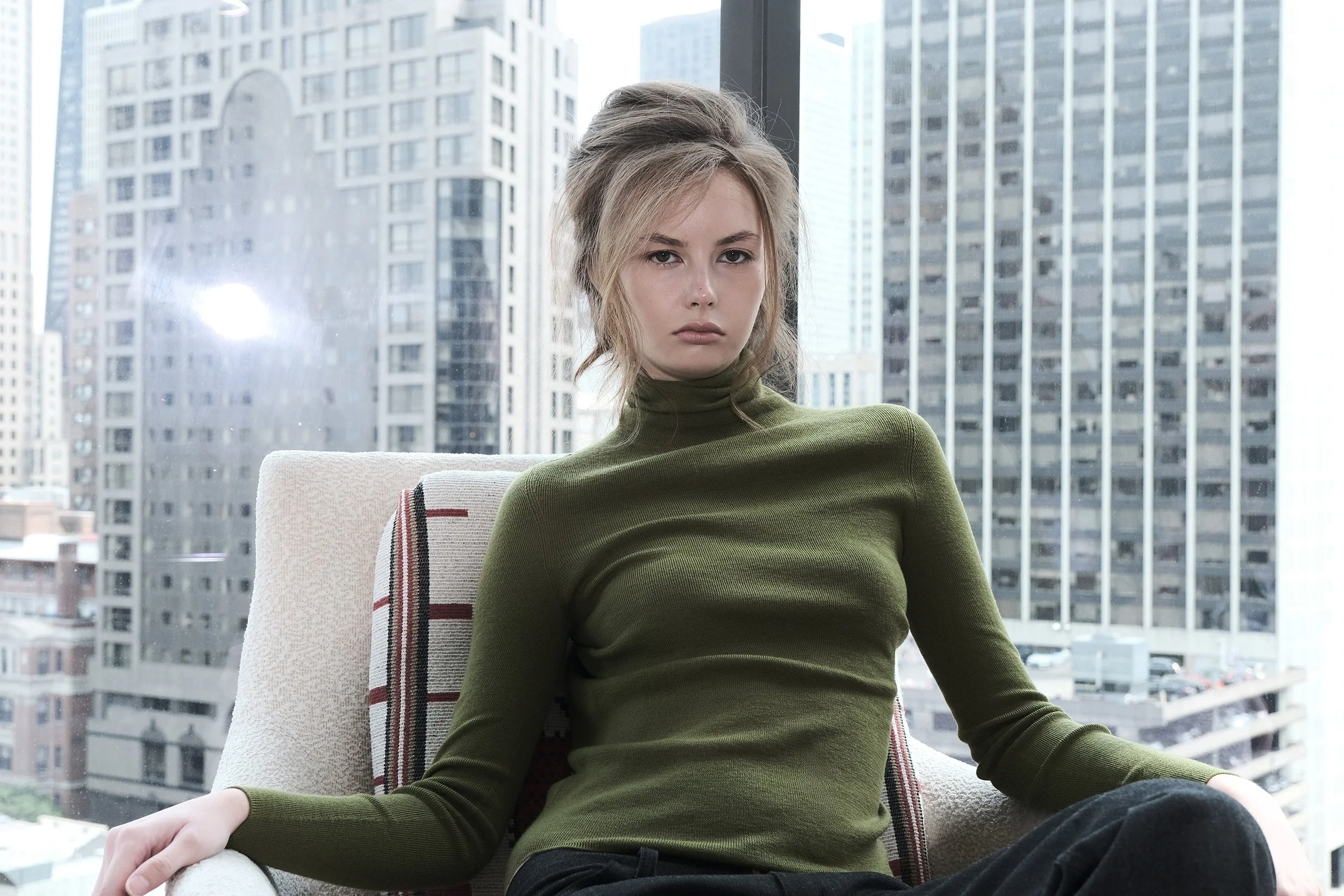 A young woman with blonde hair styled in a messy updo, wearing a green long-sleeve turtleneck, sitting in front of large windows with city buildings outside.