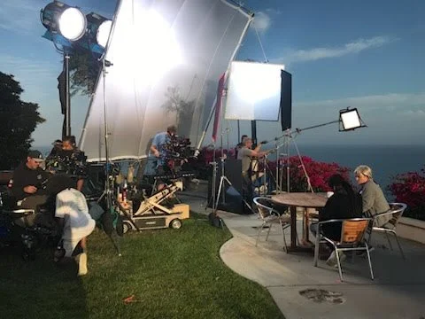 Film crew setting up equipment for filming outdoors near a coastline with pink flowers, crew members working with cameras, lights, and reflectors on a grassy and patio area.
