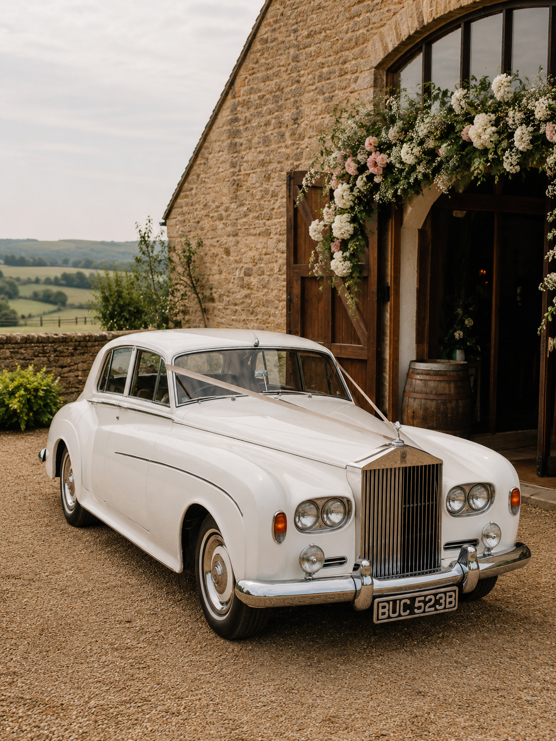 A vintage white Rolls-Royce car decorated with ribbons, parked outside a brick building with wooden doors and floral arrangements, in a rural setting with green fields in the background. Vintage wedding car for a real wedding in East Sussex