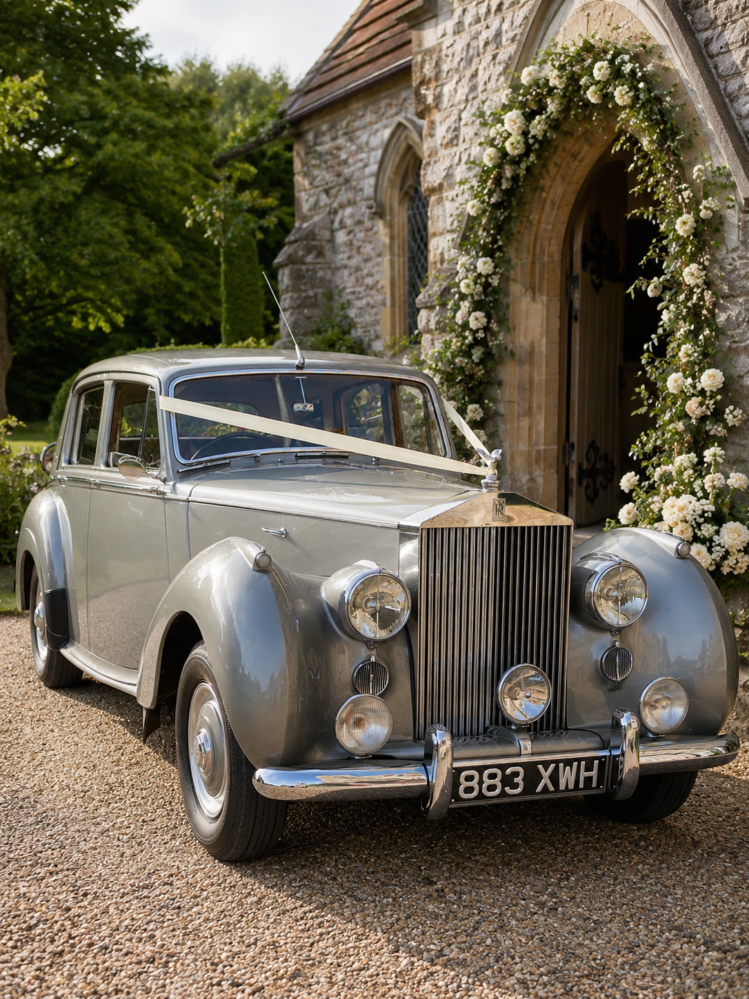 A vintage silver Rolls-Royce car decorated with a white ribbon parked in front of a stone chapel with floral arrangements around the doorway.