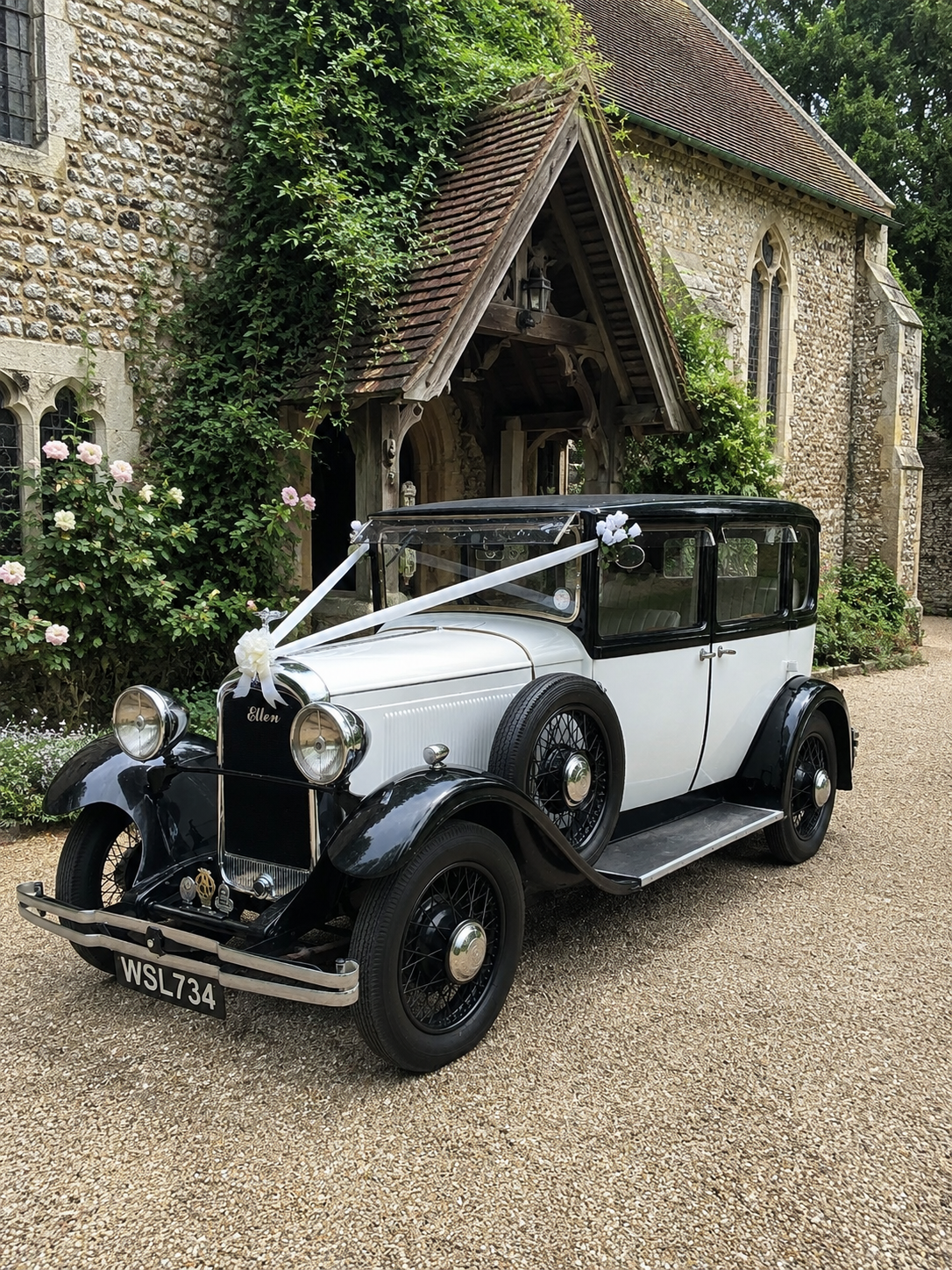 A vintage black and white car decorated with white ribbons and flowers parked in front of a stone church with ivy-covered walls and pink and white flowers.