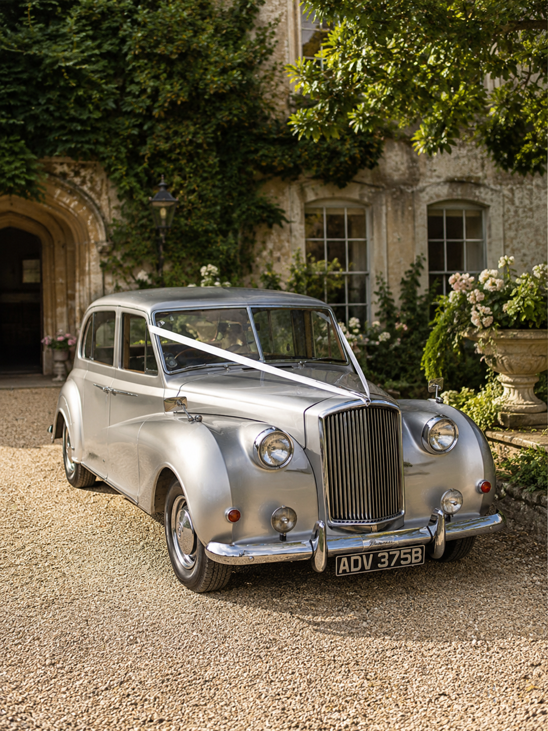 Vintage silver car decorated with a white ribbon, parked in front of an old stone building with flowering plants.