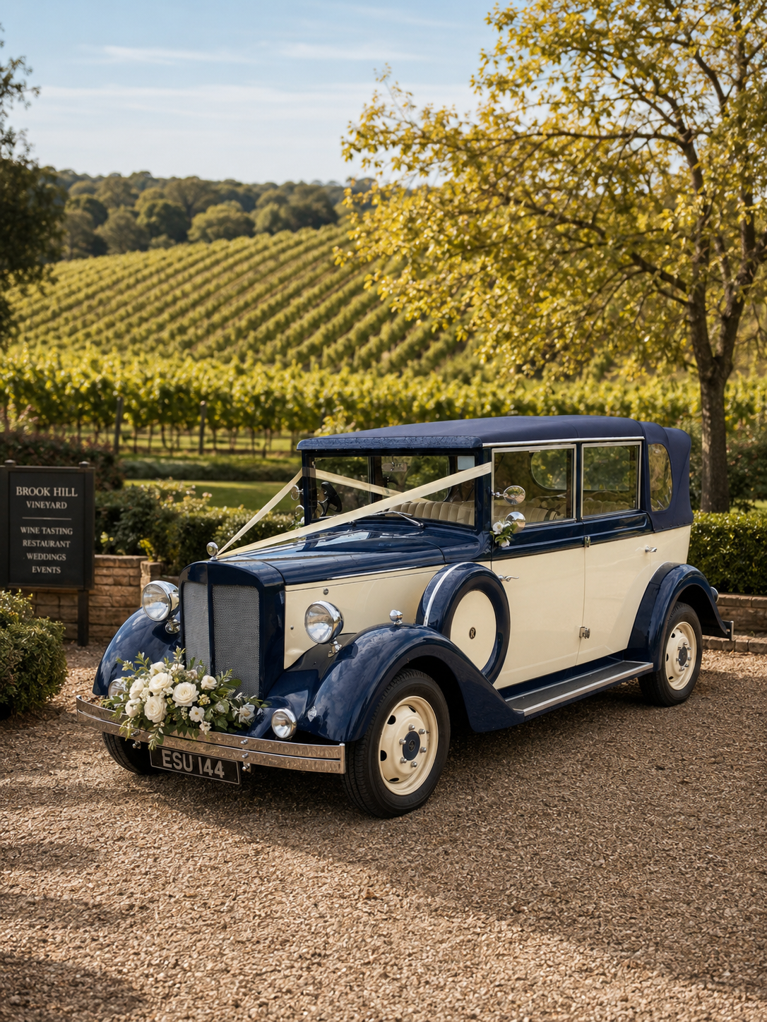 A vintage blue and cream wedding car decorated with white flowers, parked outdoors at a vineyard with rows of grapevines and a tree in the background.