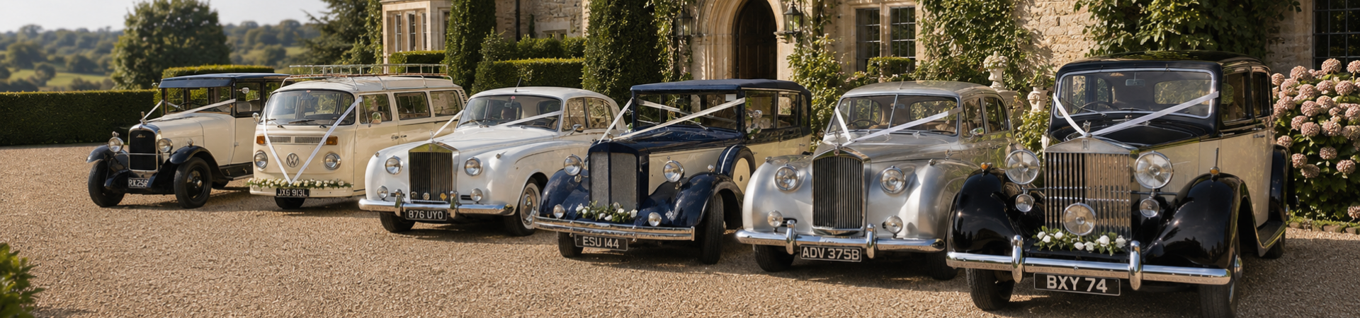 Six vintage cars decorated with white ribbons and flowers, parked in front of a stone building with flowers and greenery.