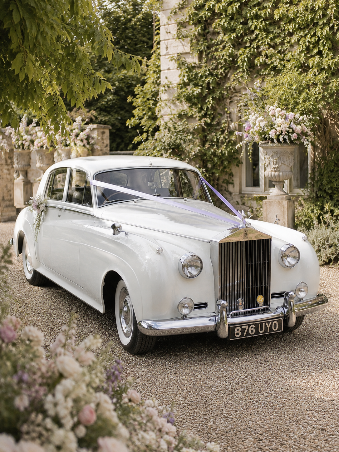 A vintage white car decorated with purple ribbons and floral arrangements parked outdoors near a stone building with greenery and flower planters.