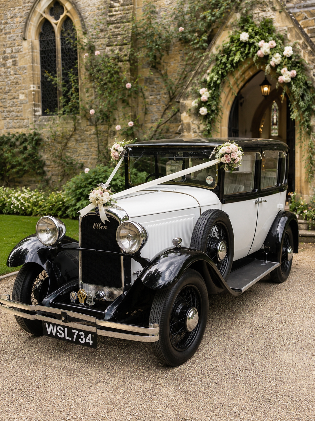 Vintage black and white car decorated with pink and white flowers for a wedding, parked outside a stone church with stained glass windows and an arched entrance.