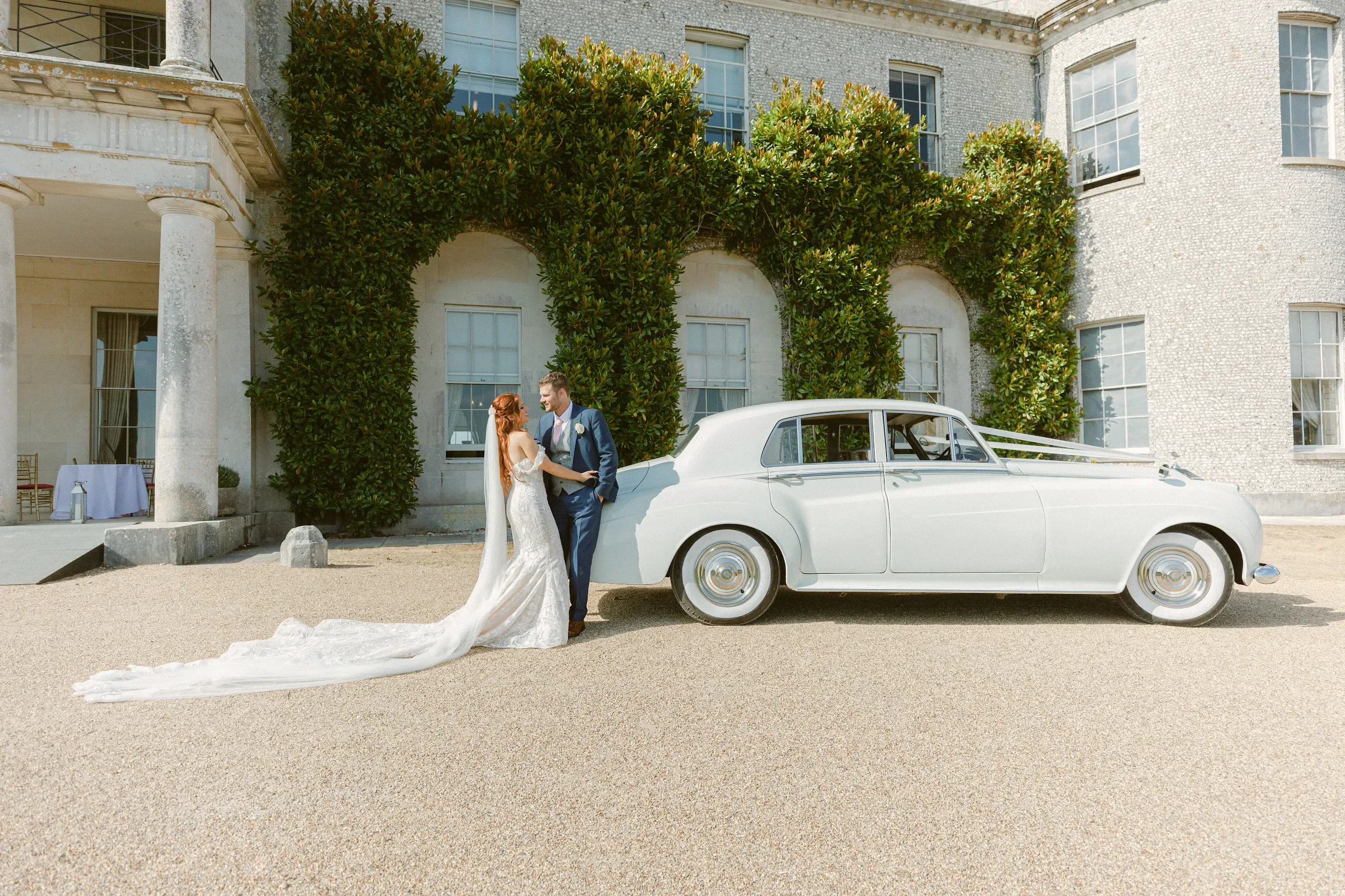 A bride and groom in wedding attire are standing outdoors near a vintage white limousine in front of a large historic building with half-timbered architecture and lush greenery.