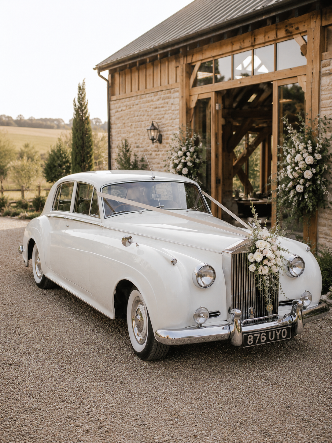 A white vintage car decorated with flower arrangements and ribbons, parked outside a rustic wooden barn decorated for a wedding.