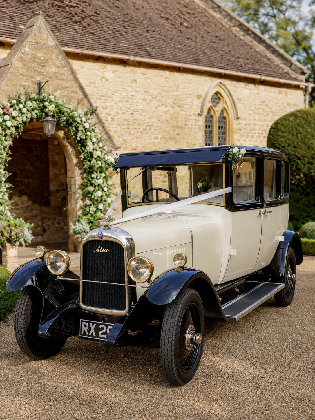 A vintage white and black car decorated with flowers and ribbons, parked outside a church with a floral arch entrance.