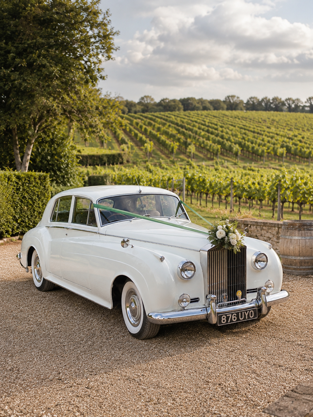 A vintage white car decorated with flowers and ribbons parked on a gravel path beside a vineyard with rolling hills and rows of grapevines in the background.