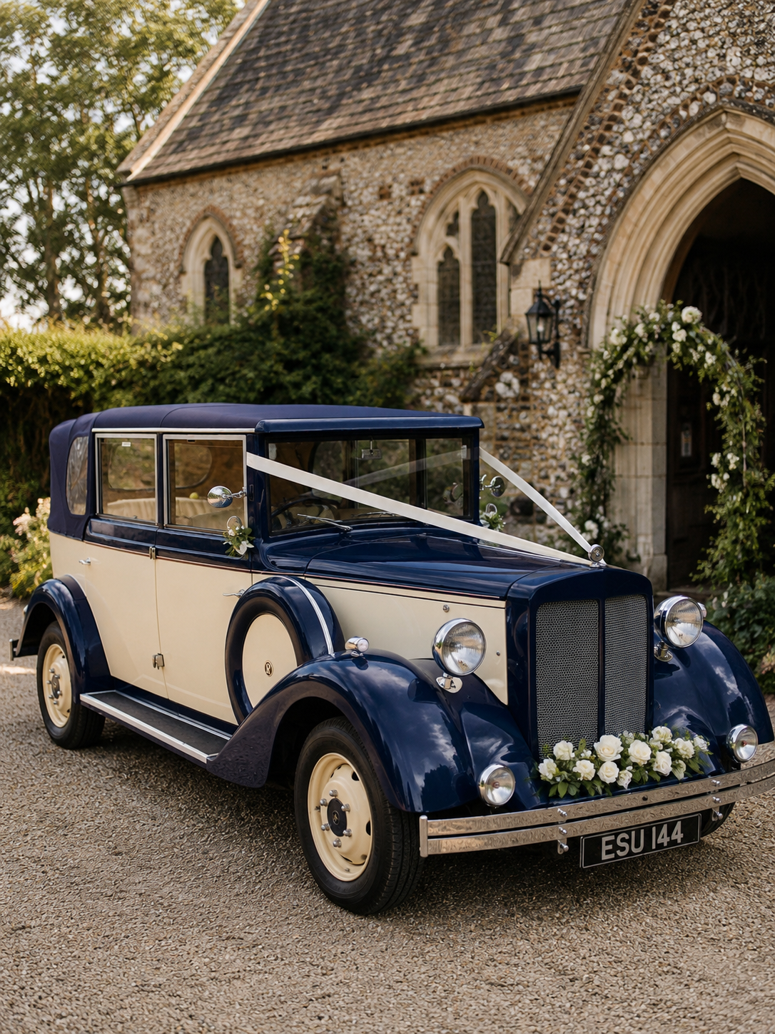 A vintage blue and cream wedding car decorated with white flowers and ribbons, parked outside a stone church with brick and stone exterior and arched doorway