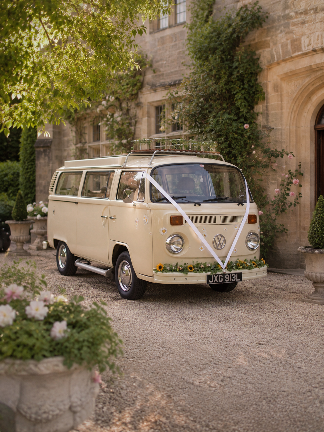 A vintage beige Volkswagen camper van decorated with flowers and a white ribbon, parked in a garden courtyard near a stone building with trees and potted plants. Vintage wedding car for a real wedding in West Sussex