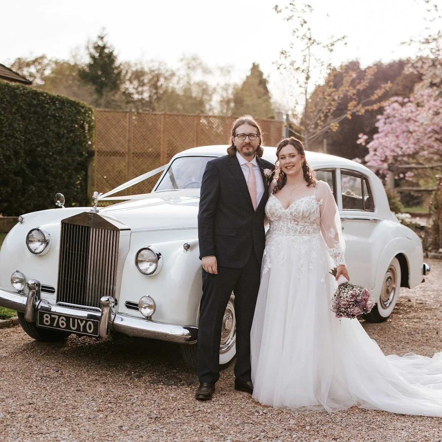 Wedding couple standing in front of a vintage white car, outdoors with trees and pink blossoms in the background.
