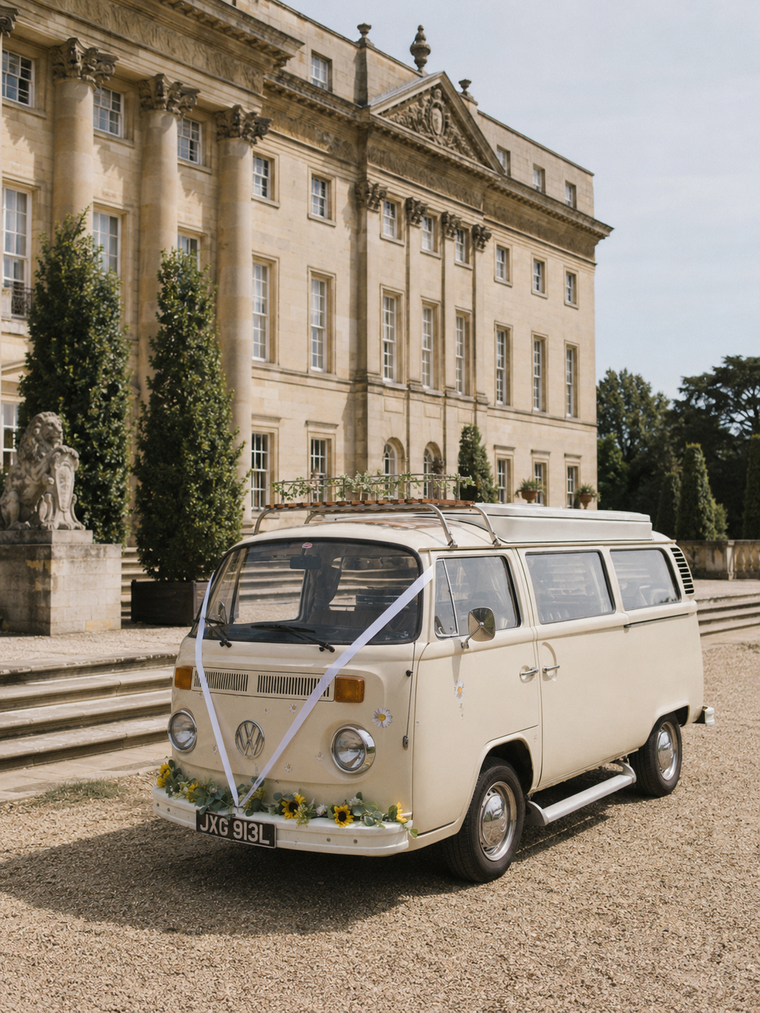 A vintage cream-colored Volkswagen van decorated with yellow sunflowers, white daisies, and wedding ribbons parked in front of a historic stone mansion with classical columns and statues. Vintage wedding car for a real wedding in West Sussex