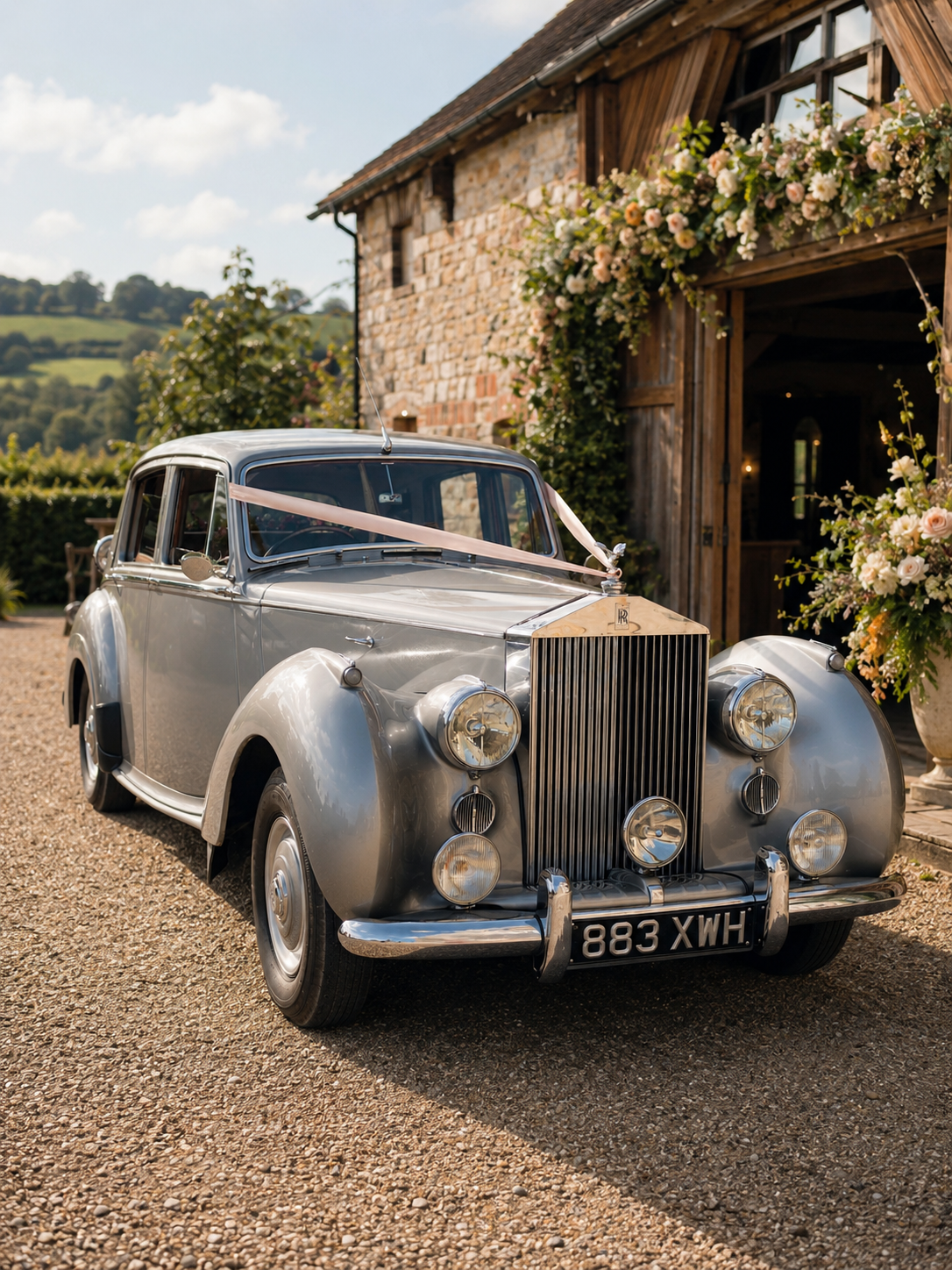 Vintage silver Rolls-Royce car decorated with a pink ribbon parked outside a rustic barn with flowers at a countryside wedding venue.