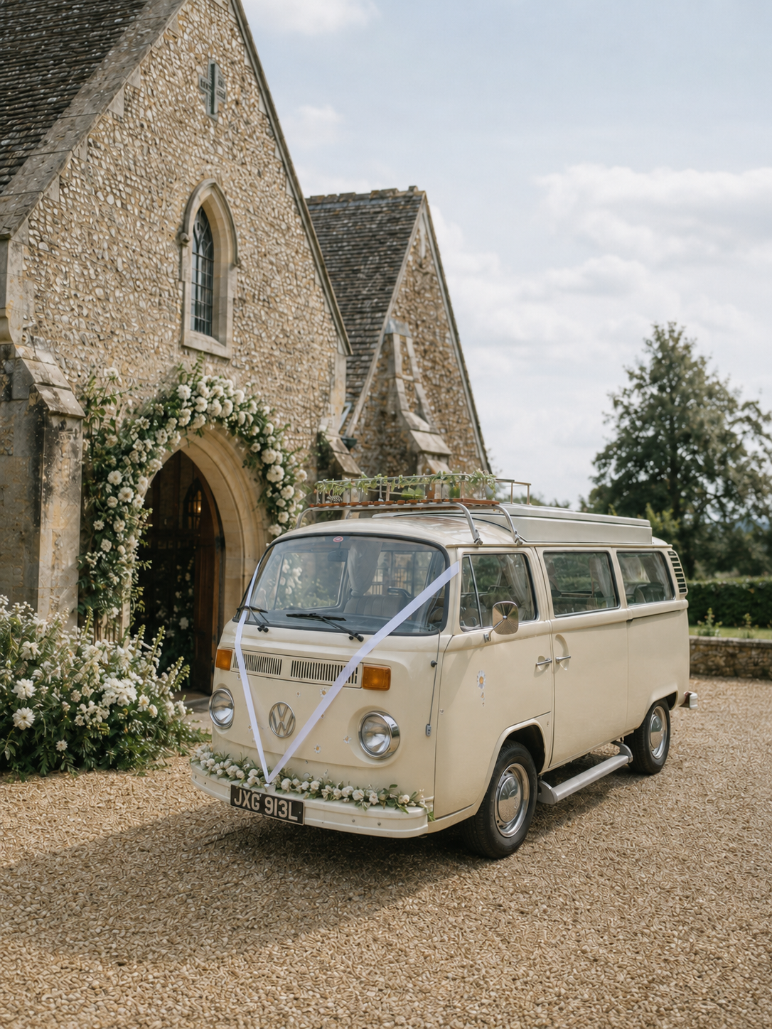 A vintage cream-colored Volkswagen van decorated with white flowers and ribbons is parked outside a stone church with an arched wooden door, surrounded by floral arrangements, on a gravel driveway under a partly cloudy sky. Vintage wedding car for a 