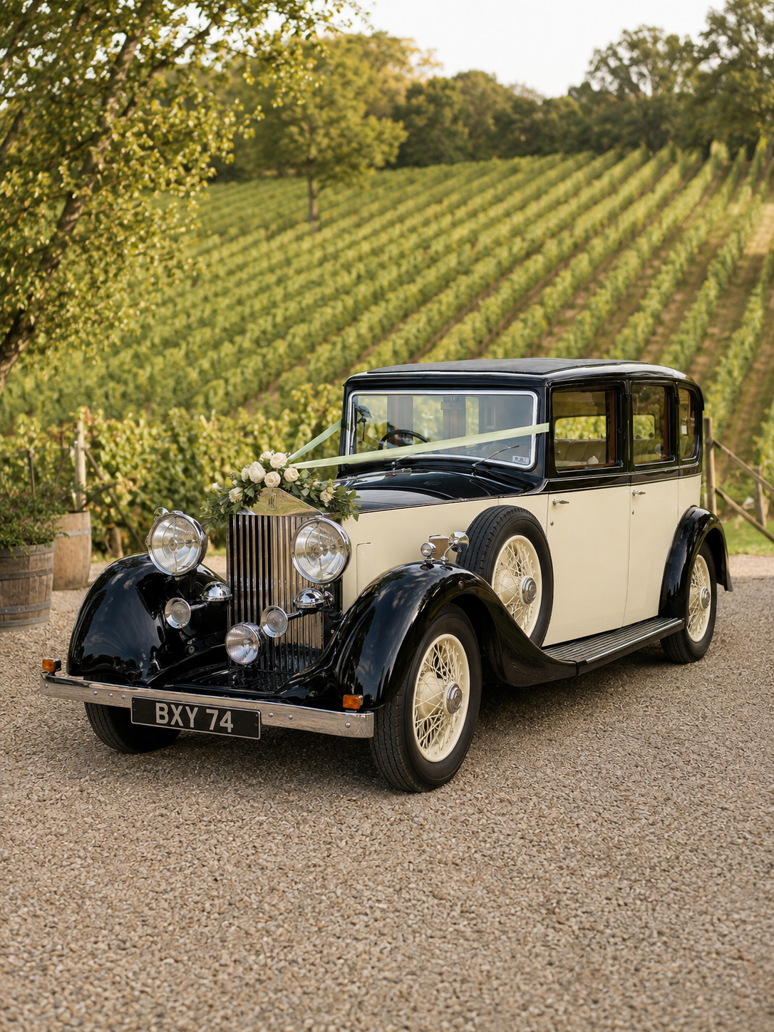 Vintage black and white car with floral decoration on the hood, parked on gravel with a vineyard in the background.