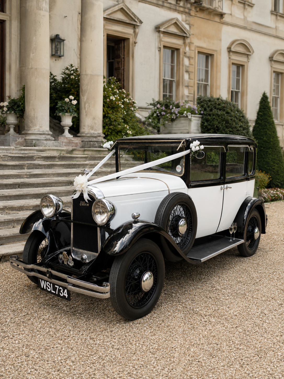 A vintage black and white car decorated with white ribbons and flowers, parked in front of a grand colonial-style building with columns, steps, and floral arrangements.