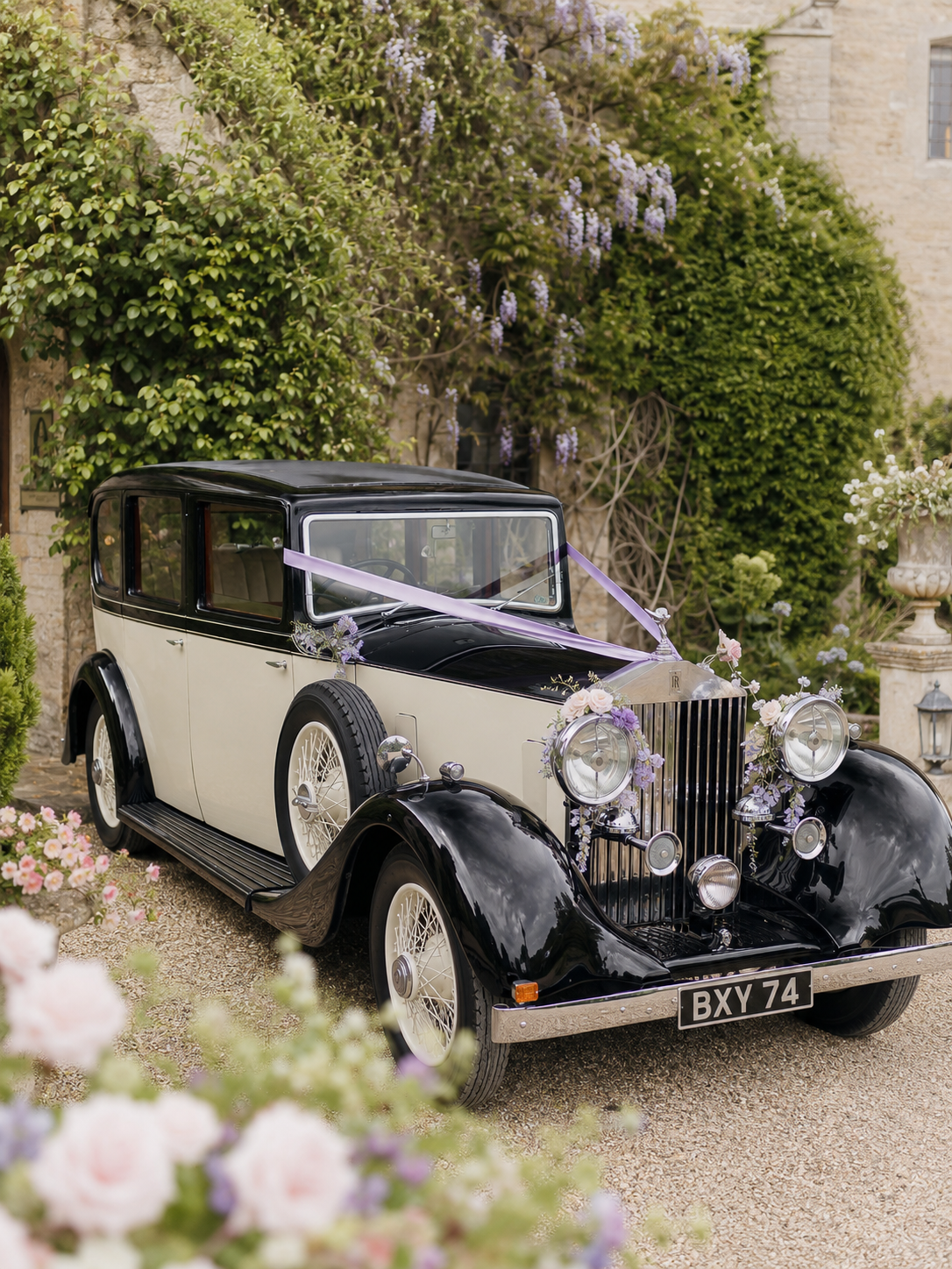 A vintage black and white car decorated with purple ribbons and flowers, parked outdoors near a stone building with lush greenery and blooming flowers.