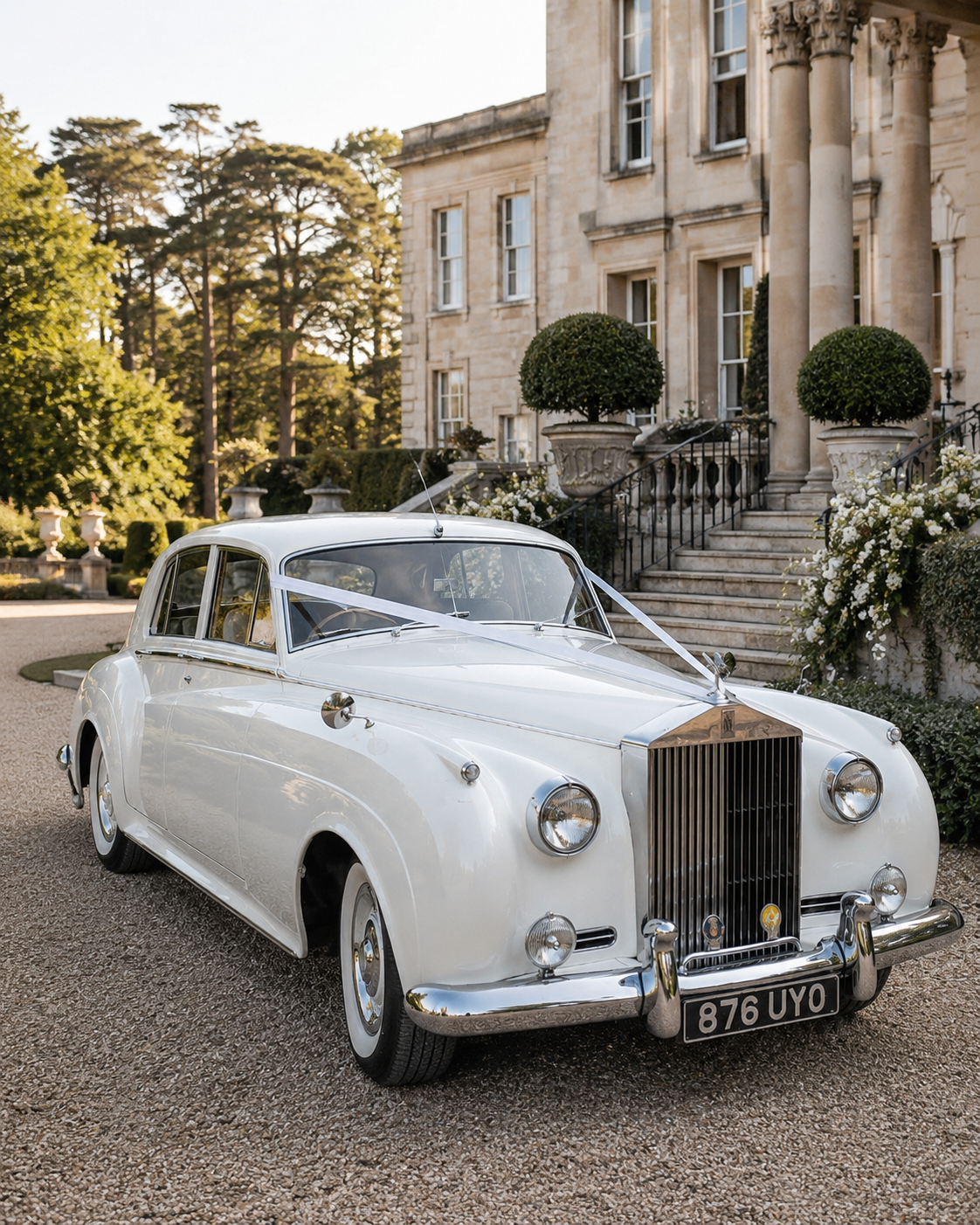 A vintage white car is parked in front of an elegant stone mansion with stairs, columns, and trimmed bushes, decorated with ribbons, at a wedding in west sussex