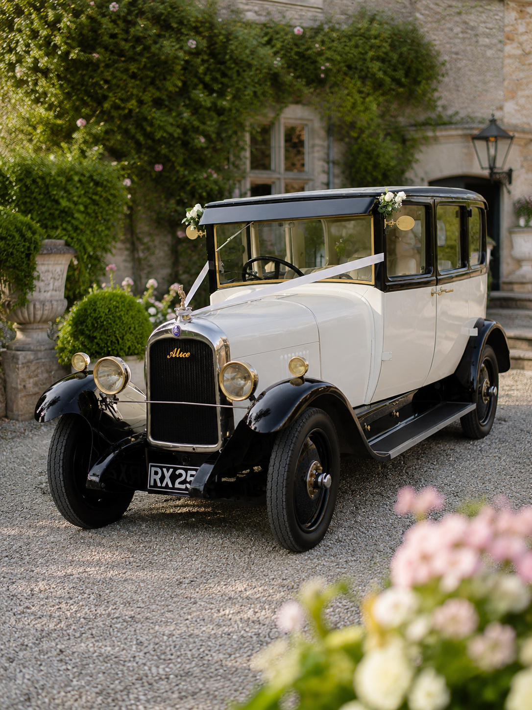 Vintage white and black car decorated with flowers and ribbons, parked on a gravel driveway in front of a stone and brick building surrounded by lush greenery and flowers.