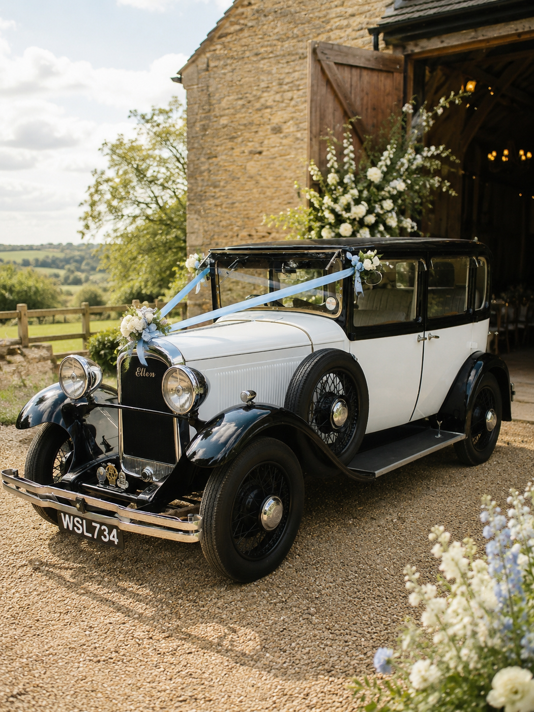 Vintage white and black car decorated with flowers and ribbons, parked outside a rustic barn for a wedding or special occasion.